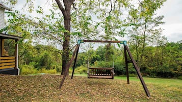 A wooden swing set with a bench, surrounded by trees and a grassy area with fallen leaves.