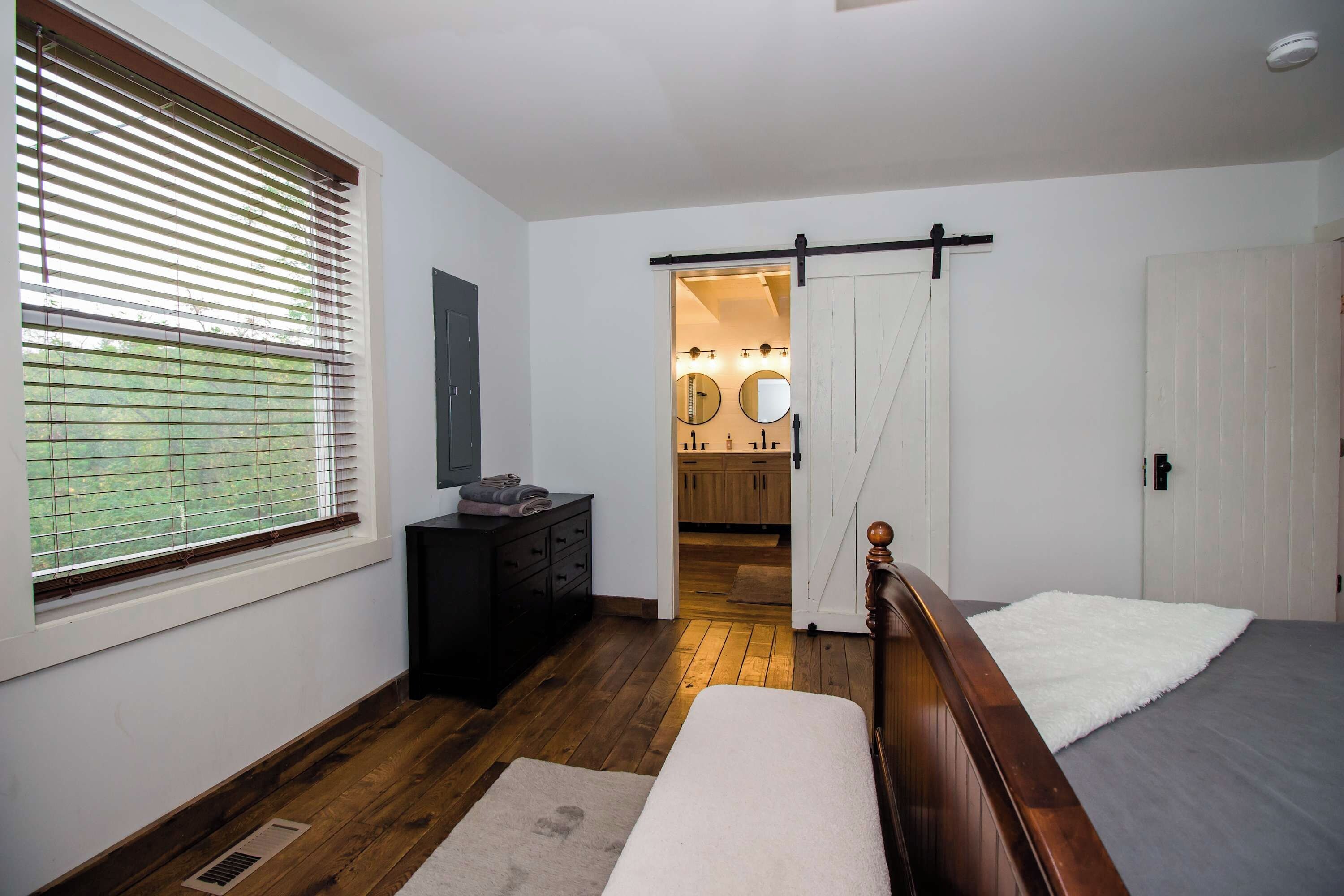 Een slaapkamer met houten vloer, een bed, een kastje en een raam met jaloezieën.