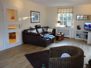 Een woonkamer met een bruine leren bank, een rieten stoel, een radiator, een televisie en ingekaderde foto's aan de wand.