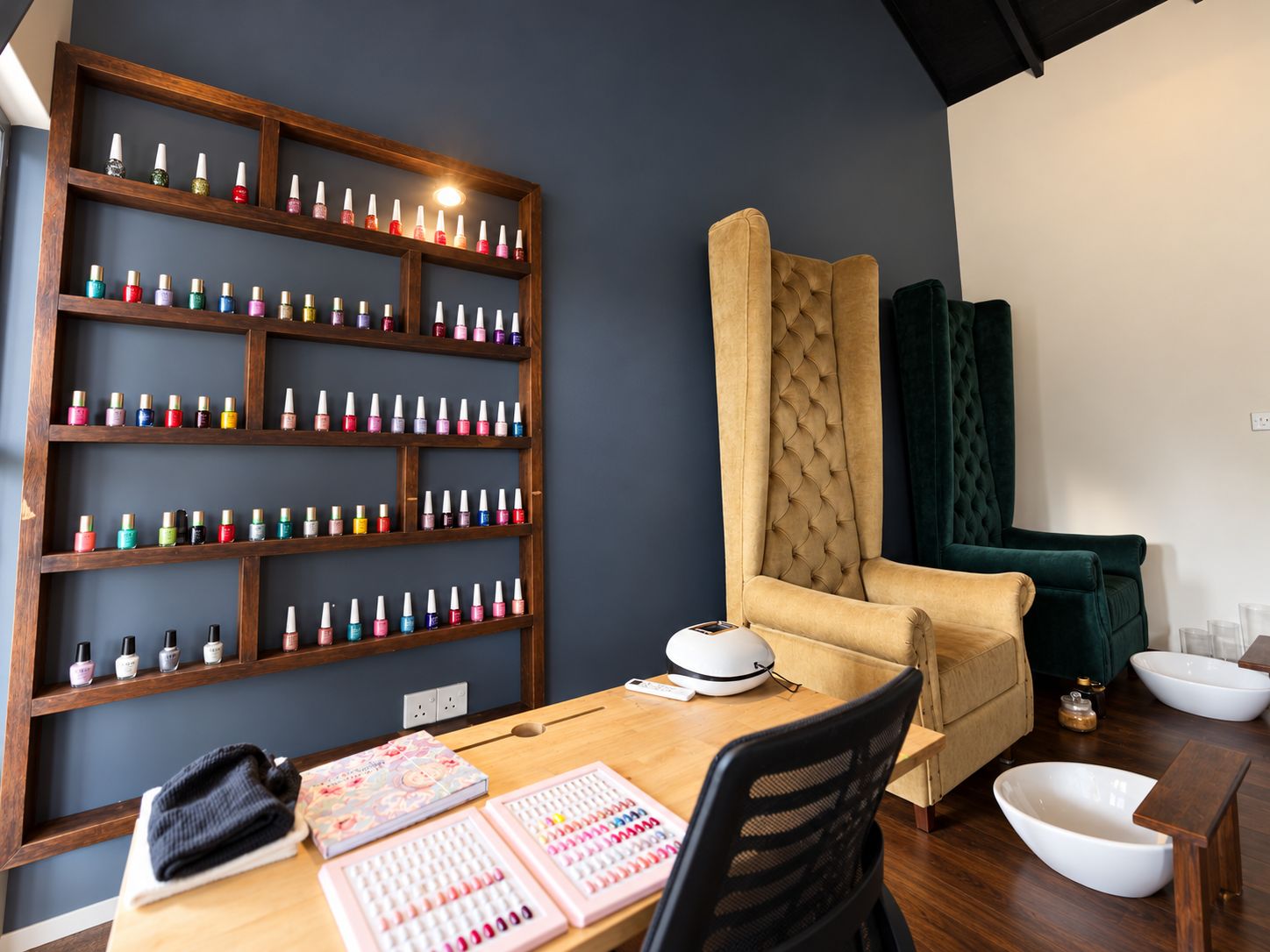A room with a wooden shelf displaying various nail polish bottles, a tufted armchair, a desk with a computer and stationery, and a small white bowl on the floor.