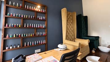 A room with a wooden shelf displaying various nail polish bottles, a tufted armchair, a desk with a computer and stationery, and a small white bowl on the floor.