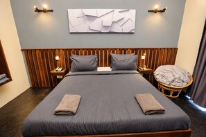 Una habitación de hotel moderna con una cama grande, cabecera de madera, dos mesitas de noche con lámparas, una obra de arte abstracto en la pared y una silla de mimbre.