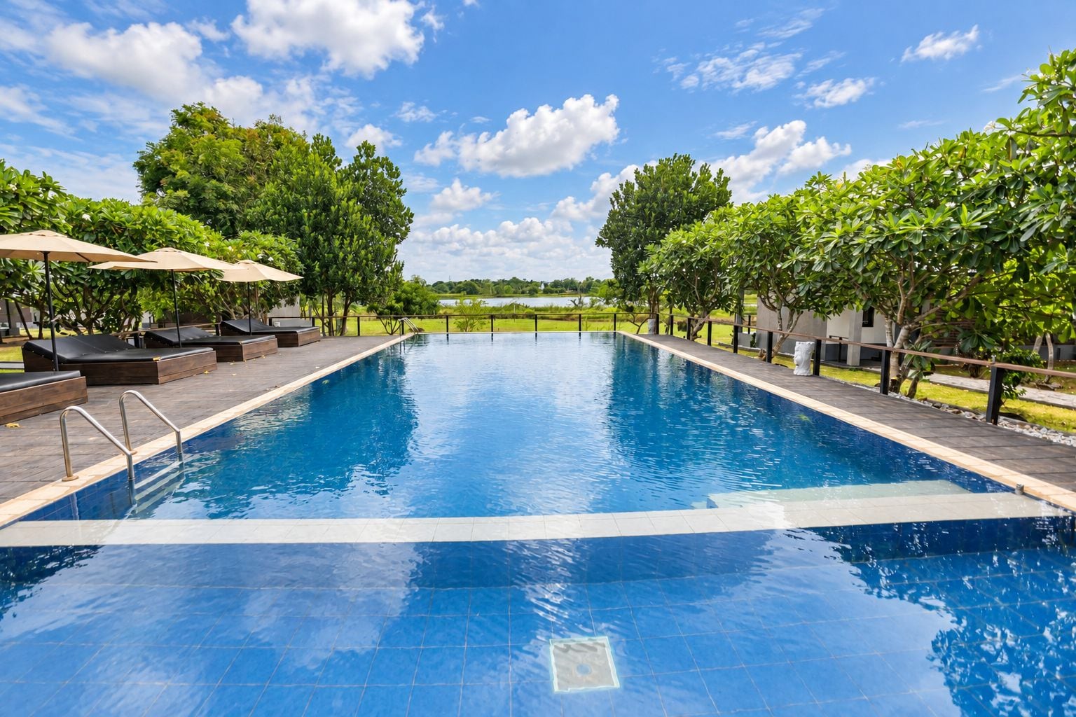 Una piscina rectangular con borde de azulejos, rodeada de sillones de descanso y sombrillas, con árboles y un cielo despejado al fondo.