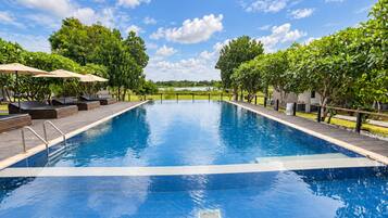 Una piscina rectangular con borde de azulejos, rodeada de sillones de descanso y sombrillas, con árboles y un cielo despejado al fondo.