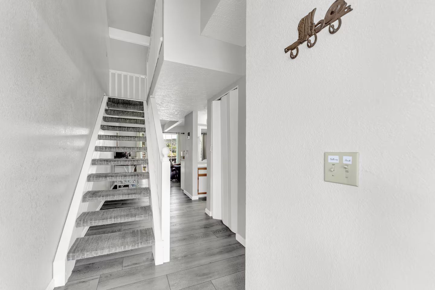 Un escalier avec des marches en bois menant à un palier, un mur blanc avec un interrupteur, et un élément décoratif en métal fixé au mur.