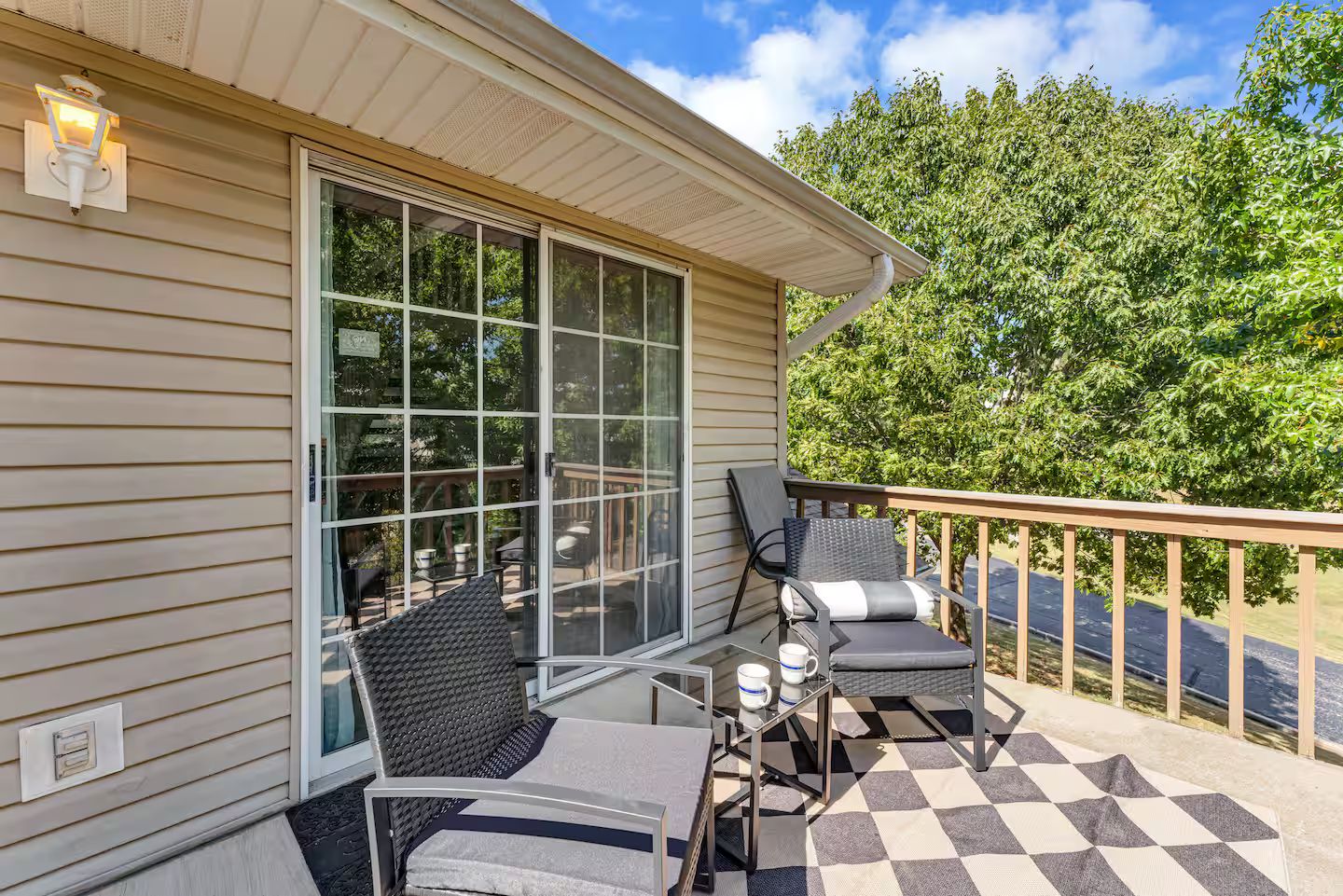 A covered outdoor area with two chairs and a table, a glass door leading to a deck, and a view of a tree-lined street.
