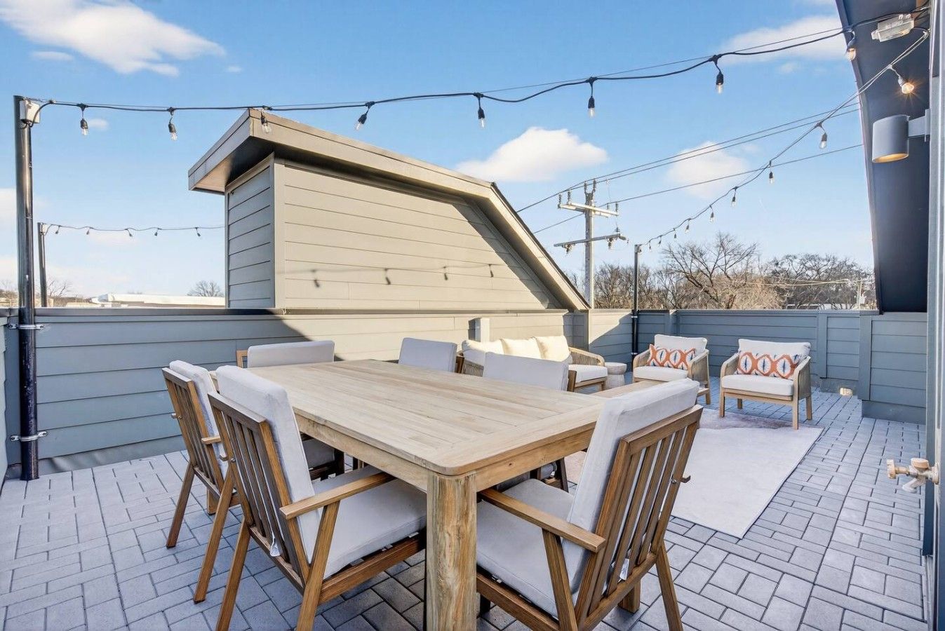 A rooftop patio with a wooden table and chairs, a small seating area with cushions, and string lights.