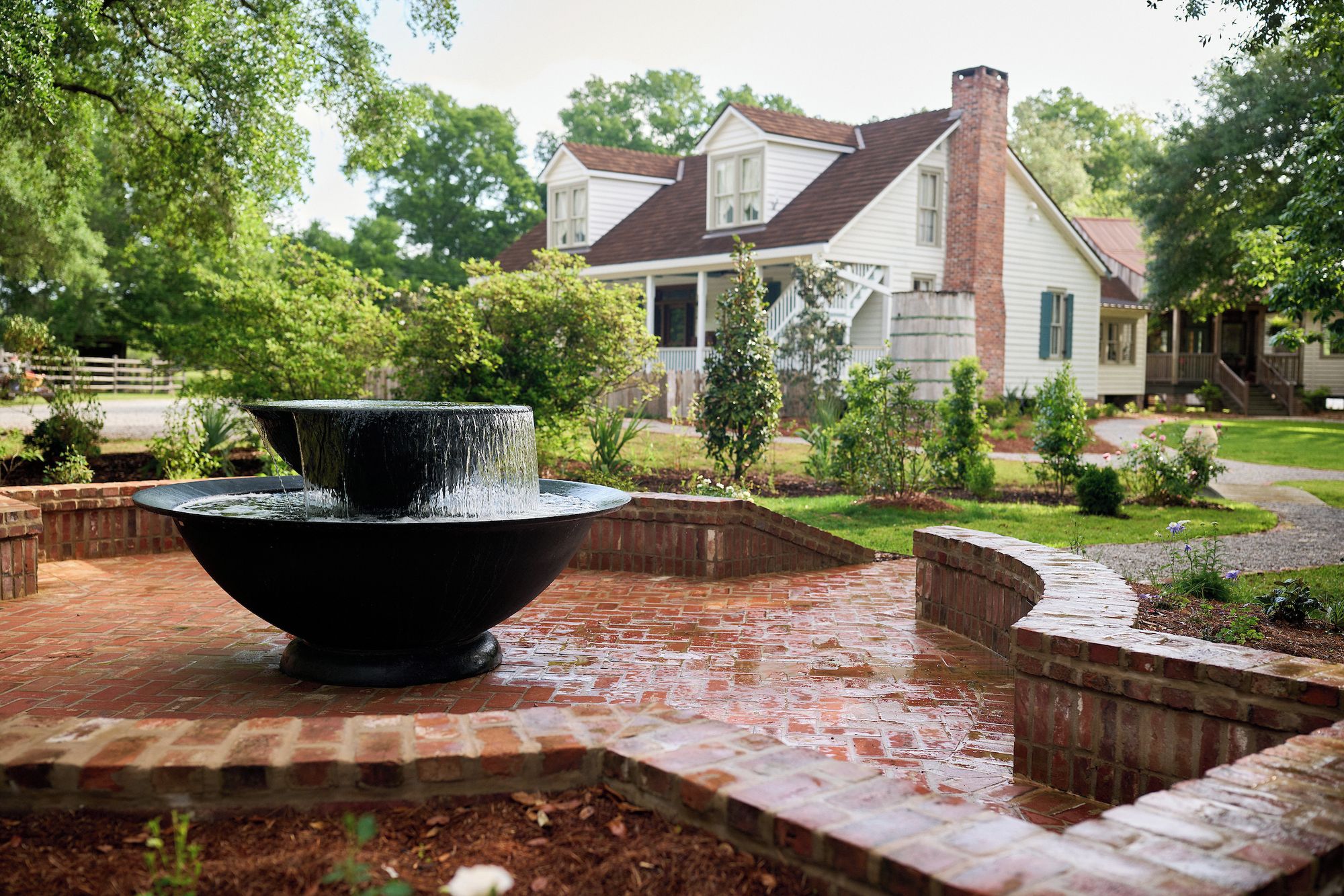 A brick patio with a black water feature in the foreground, surrounded by brick walls and greenery. In the background, there is a two-story house with a brown roof and white siding.