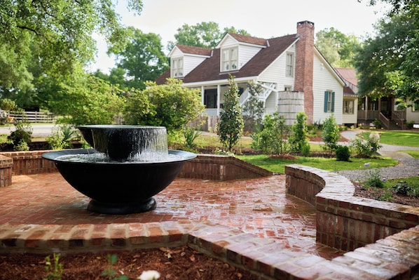 A brick patio with a black water feature in the foreground, surrounded by brick walls and greenery. In the background, there is a two-story house with a brown roof and white siding. - Family-Friendly Comfort in Acadiana: Fais-Do-Do Suite (Sunset)
