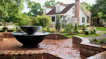 A brick patio with a black water feature in the foreground, surrounded by brick walls and greenery. In the background, there is a two-story house with a brown roof and white siding.