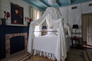 A bedroom with a four-poster canopy bed, a fireplace, and a painting on the wall.
