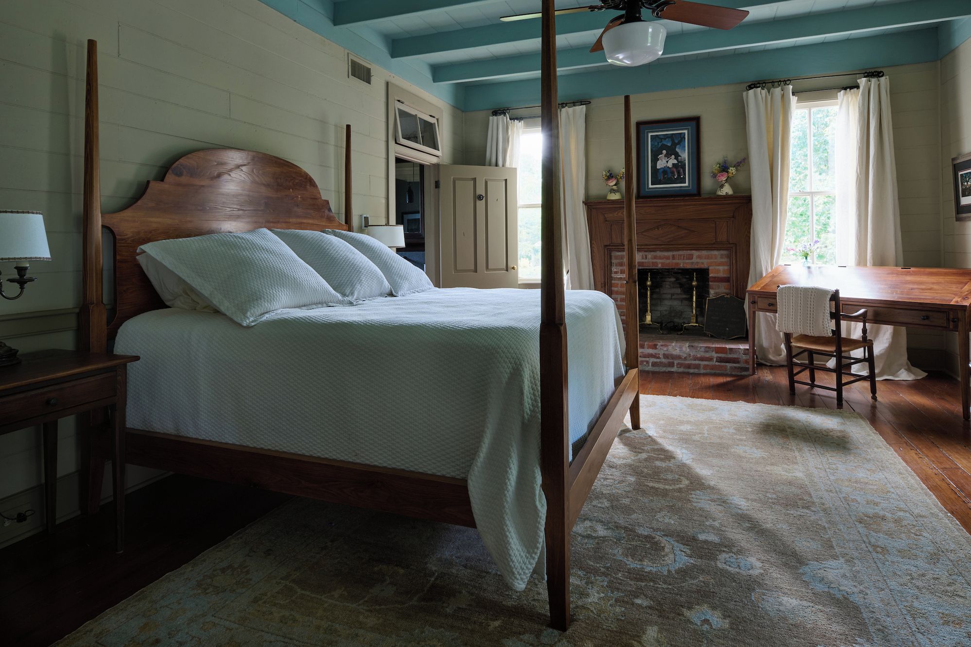 A four-poster bed with a wooden headboard, a fireplace, a dining table with chairs, and a ceiling fan.