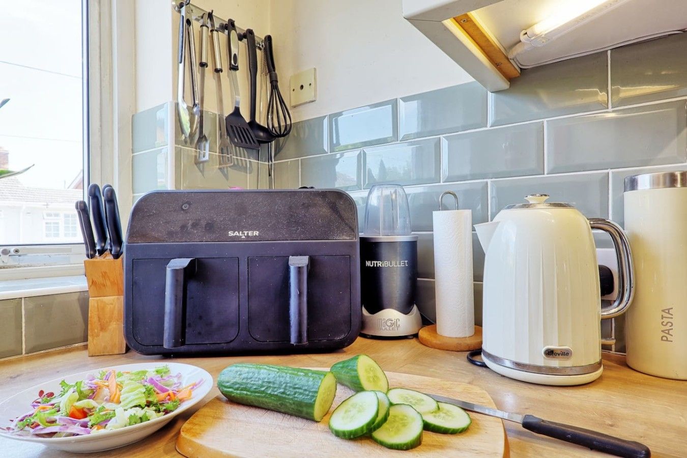 A kitchen with a food processor, a kettle, and a plate of sliced cucumbers.