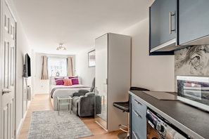 A compact hotel room with a bed, bedside table, chair, and a kitchenette area. - Host Stay Poplar Apartment (Rochester)