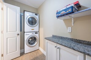 A laundry room with a stacked washer and dryer, a countertop, and a shelf with board games. - Ground-Floor Golf Course Retreat (Miramar Beach)