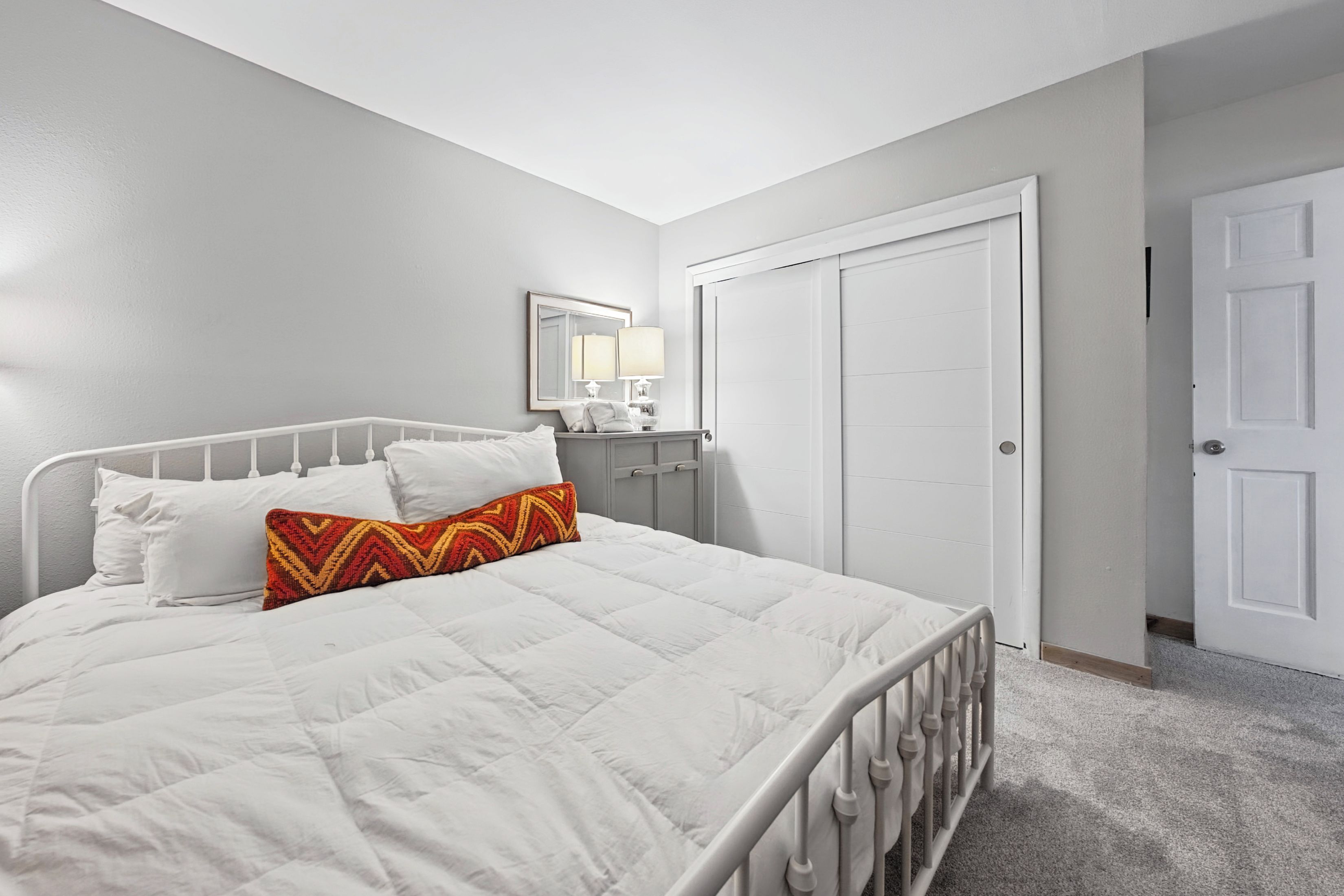 A bedroom with a white bed, a nightstand, a mirror, and a door.