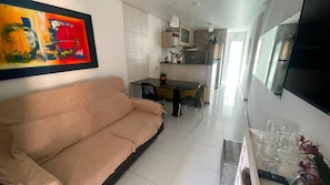 A living room with a sofa, a dining table with chairs, a kitchen area with cabinets and appliances, and a TV mounted on the wall. - Porto Paradise, 300 meters from the sea. (Ipojuca)