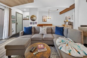 A modern living room with a sofa, ottoman, and a coffee table. There are wall decorations, a chandelier, and a kitchen area visible. - Meadow Ridge Court 22 Unit 15 clubhouse, private deck, views (Fraser)