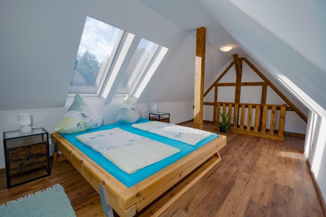 Un dormitorio con techo inclinado, vigas de madera, una cama con ropa de cama azul y tragaluces.
