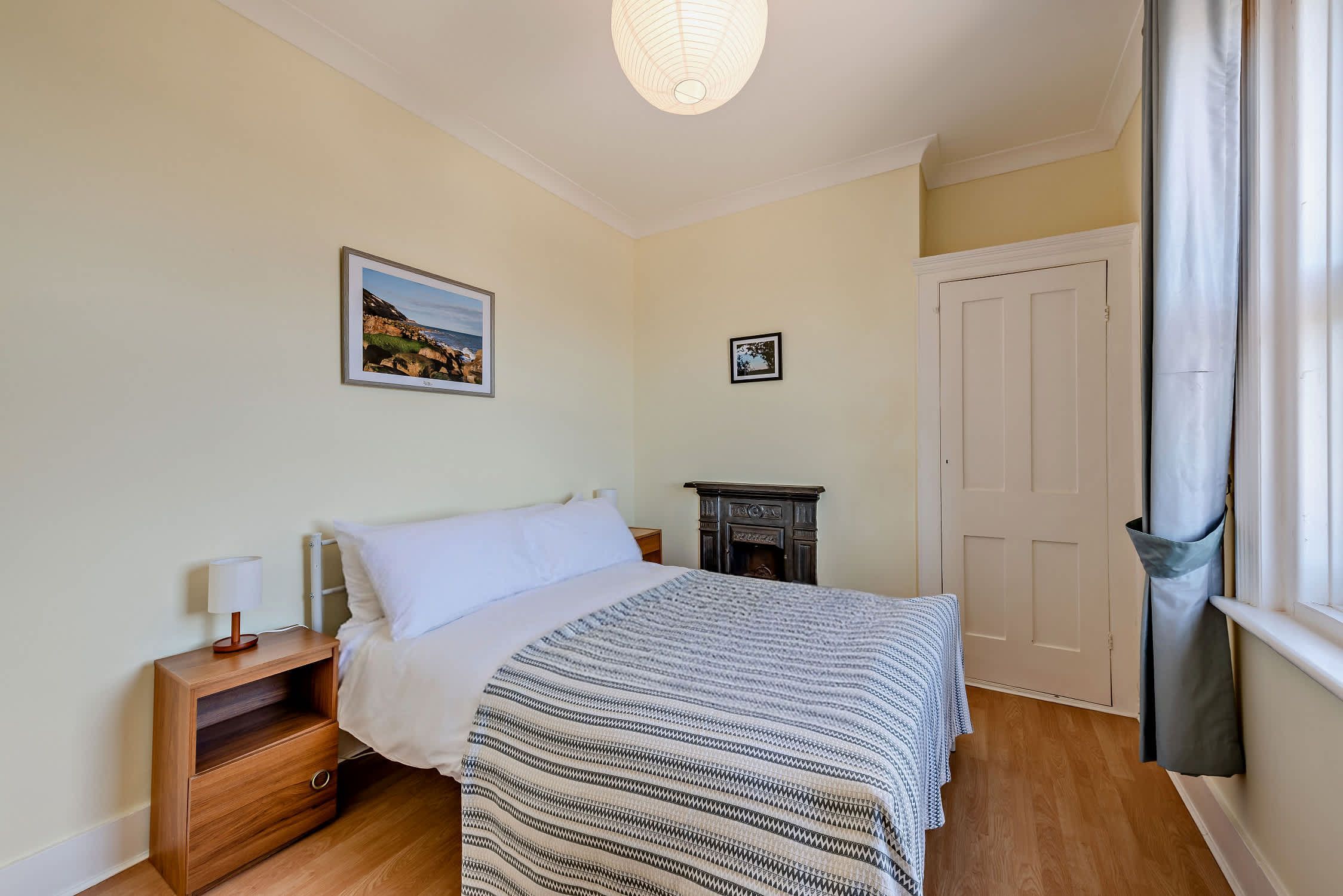 A bedroom with a bed, bedside table, lamp, framed pictures, a fireplace, and a door.