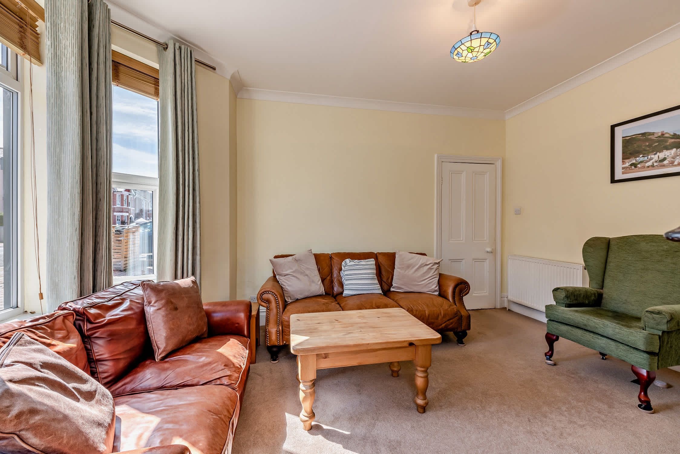 A living room with a brown leather sofa, a wooden coffee table, and a green armchair. There is a door and a framed picture on the wall.