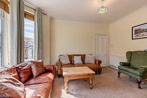 A living room with a brown leather sofa, a wooden coffee table, and a green armchair. There is a door and a framed picture on the wall.