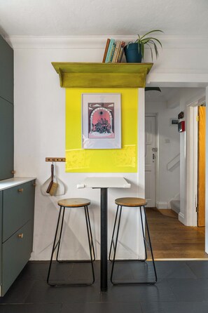Un coin repas avec un mur d’accent jaune, un tableau encadré et deux tabourets.