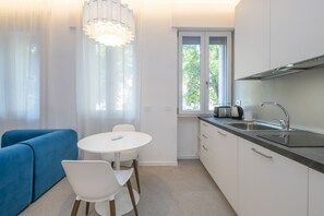 A compact kitchen with a dining area, a blue sofa, and a window with a view of greenery. - Brand-New Modern Studio, 10-Minute Walk from Bocconi (Milan)