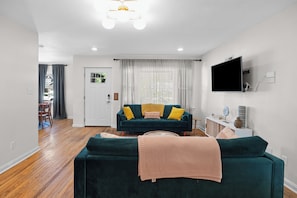 A living room with a dark green sofa, a flat-screen TV mounted on the wall, and a wooden floor. - Campus Edge Cottage - Serene 3-bedroom house in West Lafayette (West Lafayette)