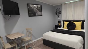 Un dormitorio con cama, escritorio, silla, televisor montado en la pared y un atrapasueños.