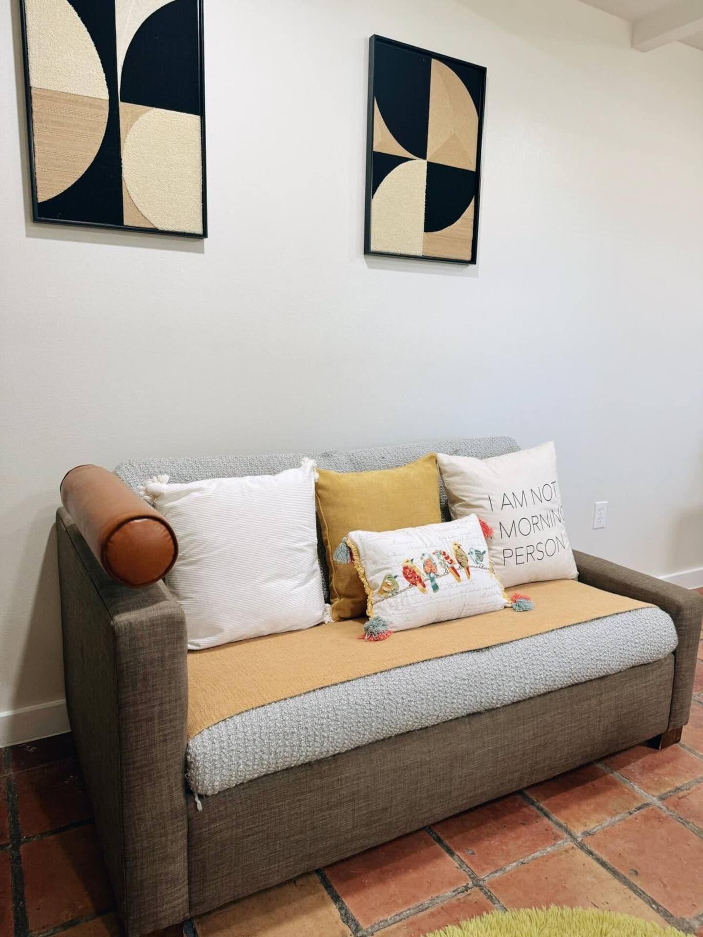 Un canapé gris avec un coussin marron, deux oreillers et un plaid dans une pièce dont le sol est carrelé et les murs sont blancs.