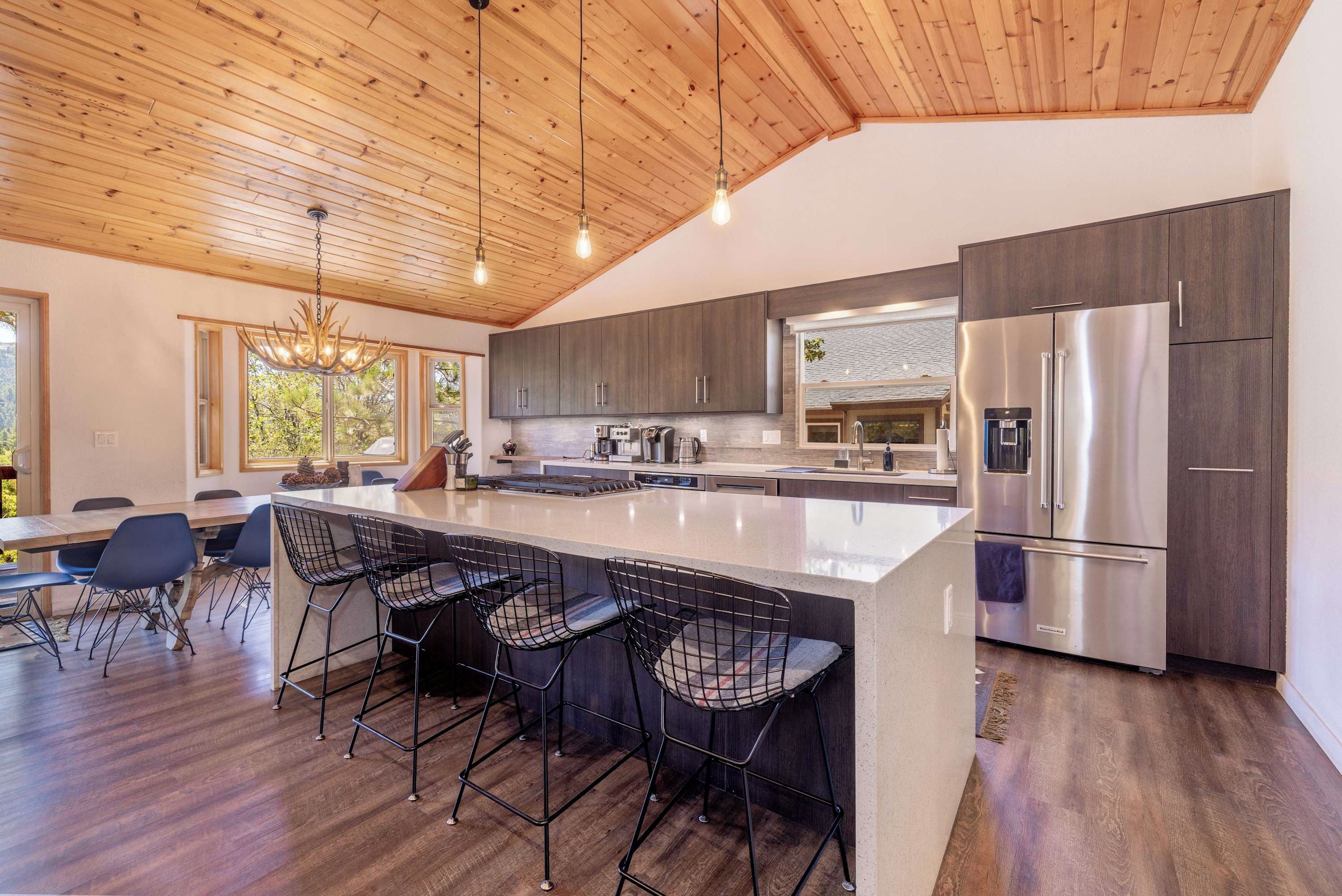 A modern kitchen with a large island, bar stools, stainless steel appliances, and a wooden ceiling.