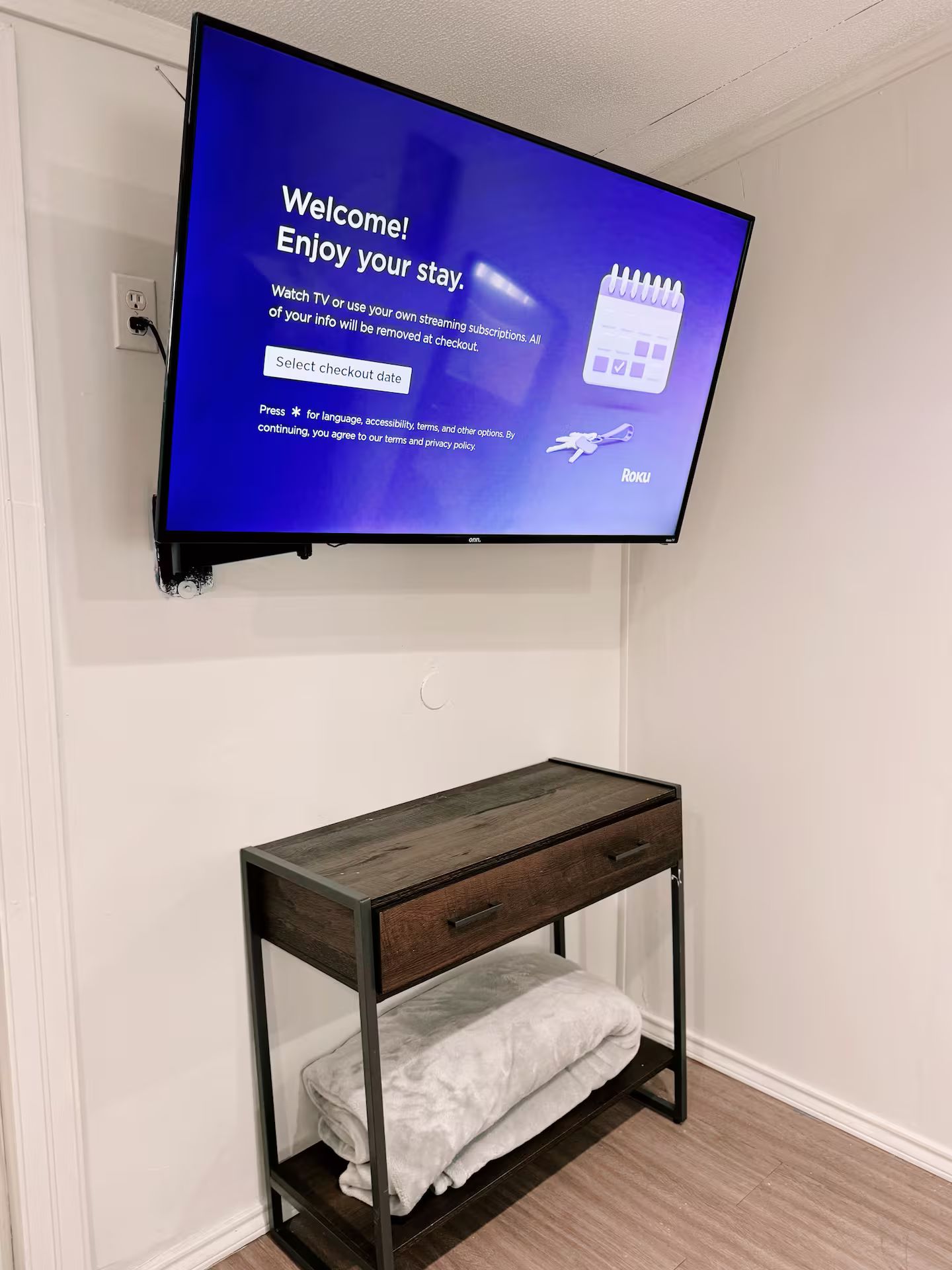 A television mounted on a wall displaying a welcome message and checkout options. Below the TV, there is a wooden bench with a cushion.