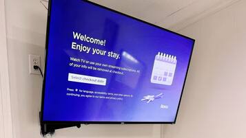 A television mounted on a wall displaying a welcome message and checkout options. Below the TV, there is a wooden bench with a cushion.