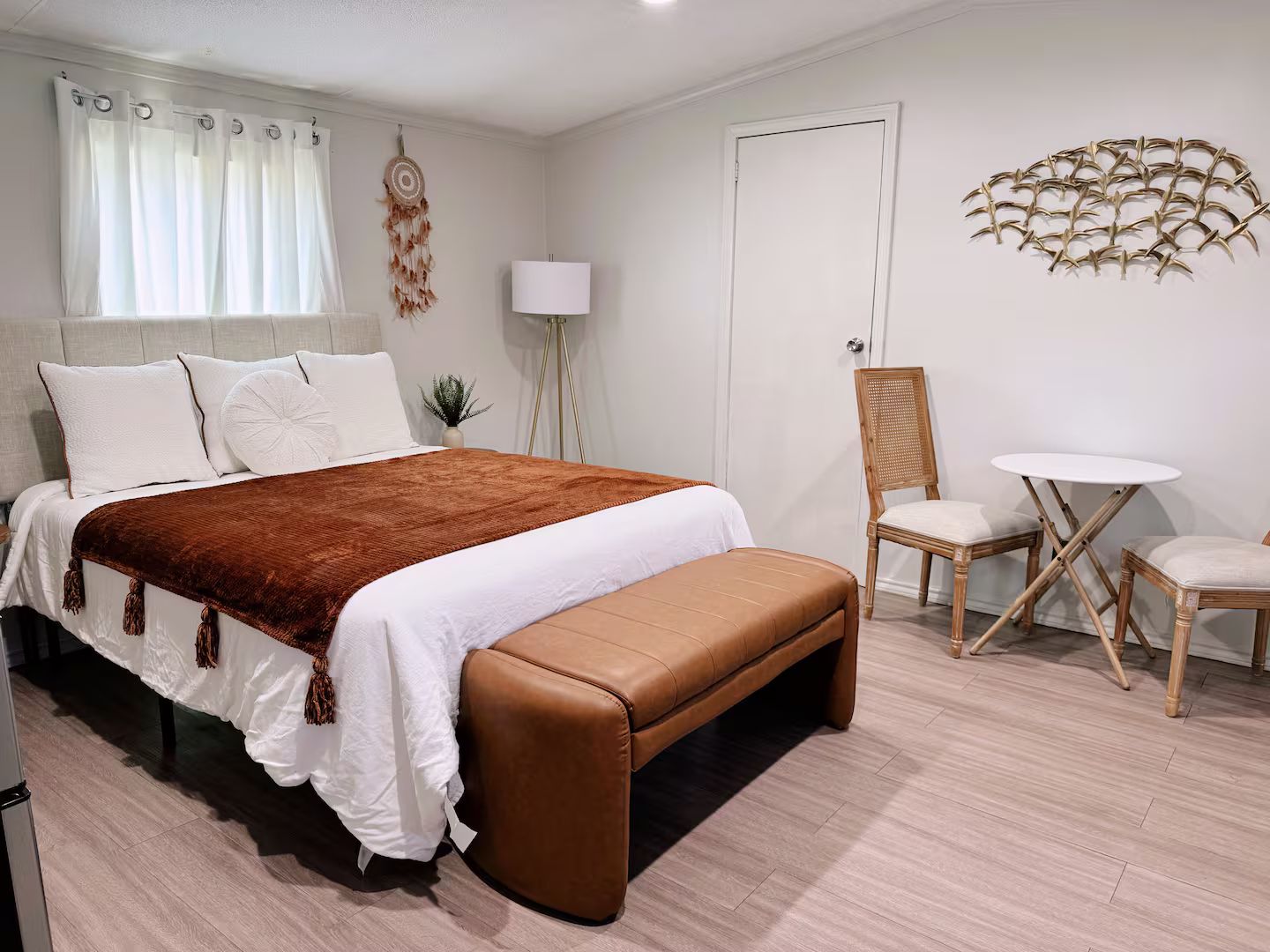 Quarto com cama, um banquinho, uma cadeira, uma mesinha e um objeto decorativo na parede.