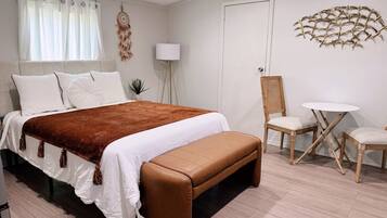 Quarto com cama, um banquinho, uma cadeira, uma mesinha e um objeto decorativo na parede.