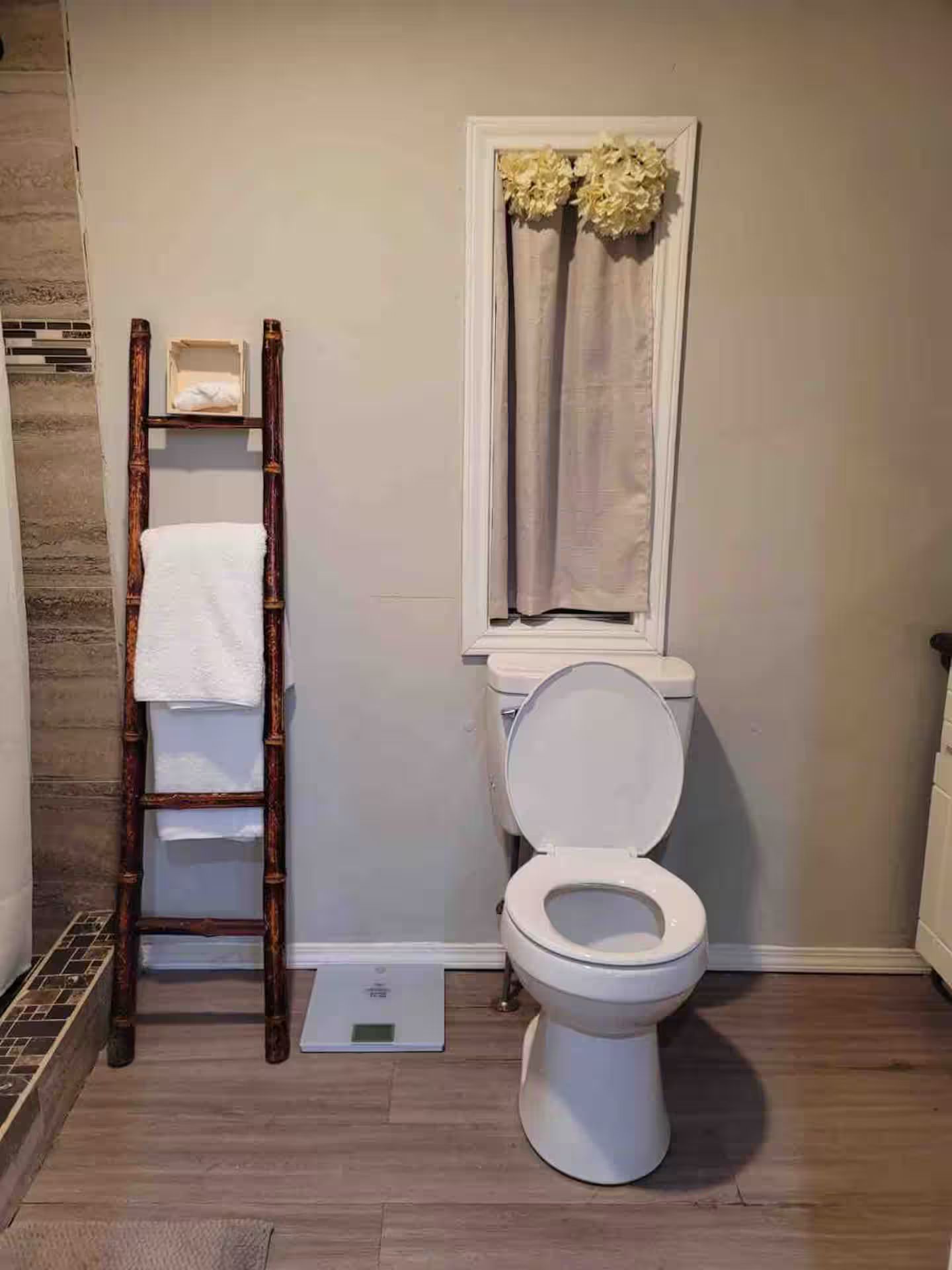 Ein Badezimmer mit Toilette, einer Holzleiter, einer Waage und einem Fenster mit Vorhängen.