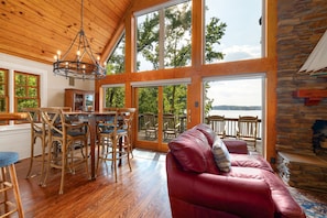 A spacious room with a wooden ceiling, a dining area with a table and chairs, a red leather sofa, and a large window overlooking a body of water. - Summer Place A-Frame • Luxury Main Lake A-Frame • Private Beach + Sleeps 10 (Littleton)