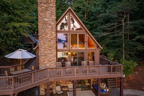 A two-story cabin with a stone chimney, wooden deck, and glass doors leading to the interior. - Summer Place A-Frame • Luxury Main Lake A-Frame • Private Beach + Sleeps 10 (Littleton)
