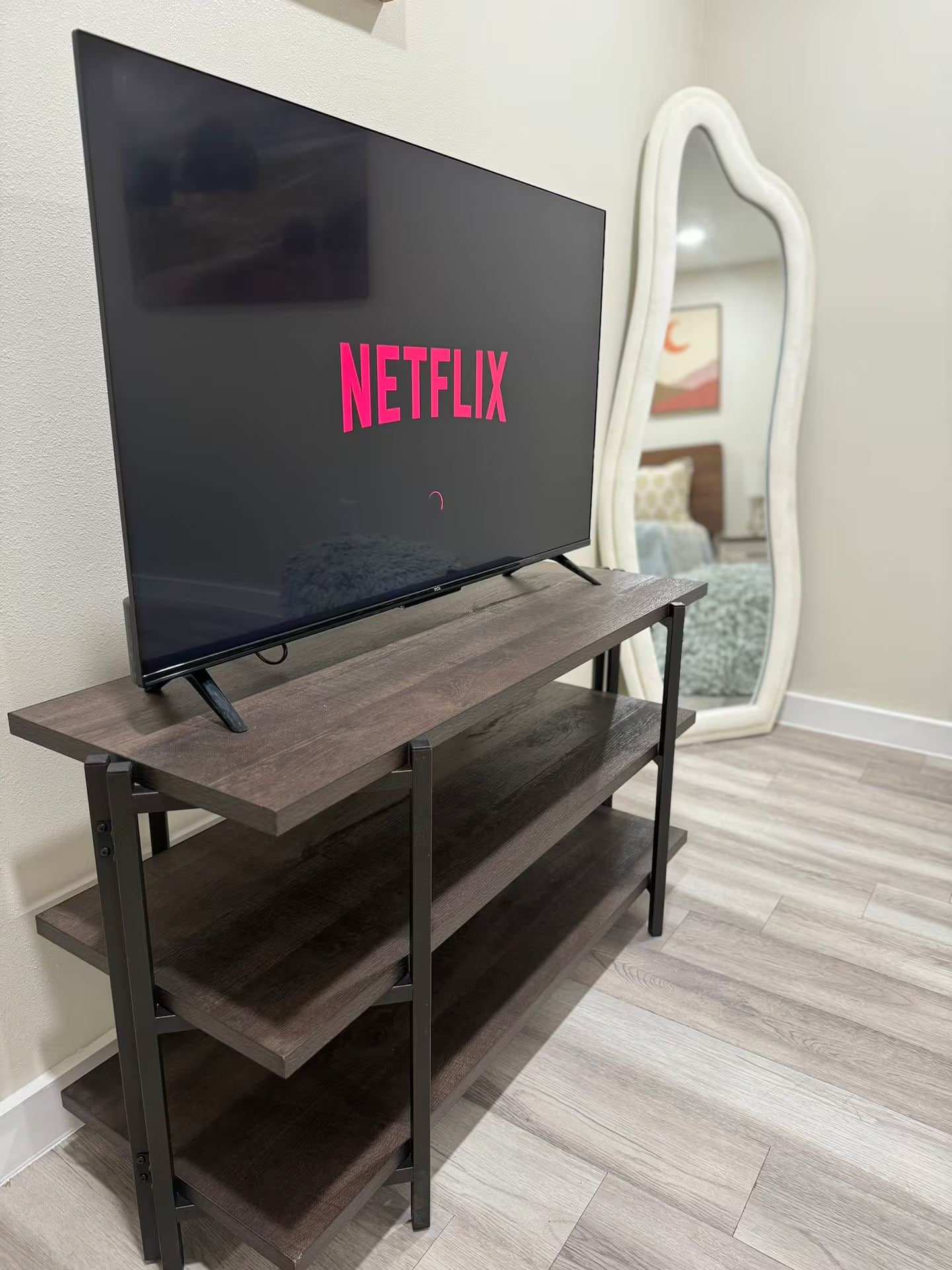 Un téléviseur à écran plat posé sur un meuble TV en bois, doté d’une étagère, dans une pièce où l’on aperçoit un miroir et un lit au fond.
