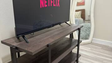 Un téléviseur à écran plat posé sur un meuble TV en bois, doté d’une étagère, dans une pièce où l’on aperçoit un miroir et un lit au fond.