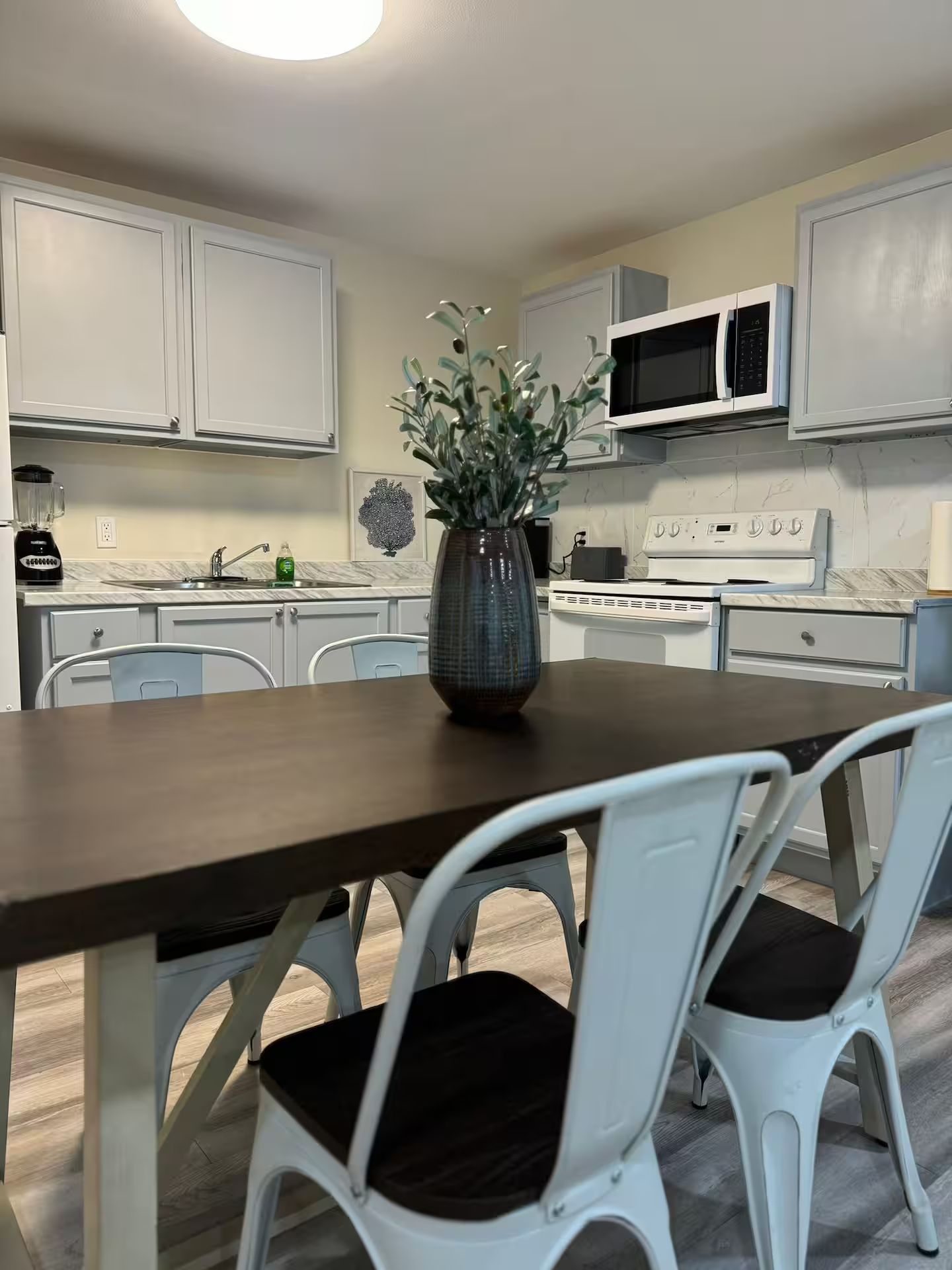 Une cuisine avec des meubles blancs, un four à micro-ondes, une cuisinière, une table à manger avec deux chaises et un vase avec des plantes.