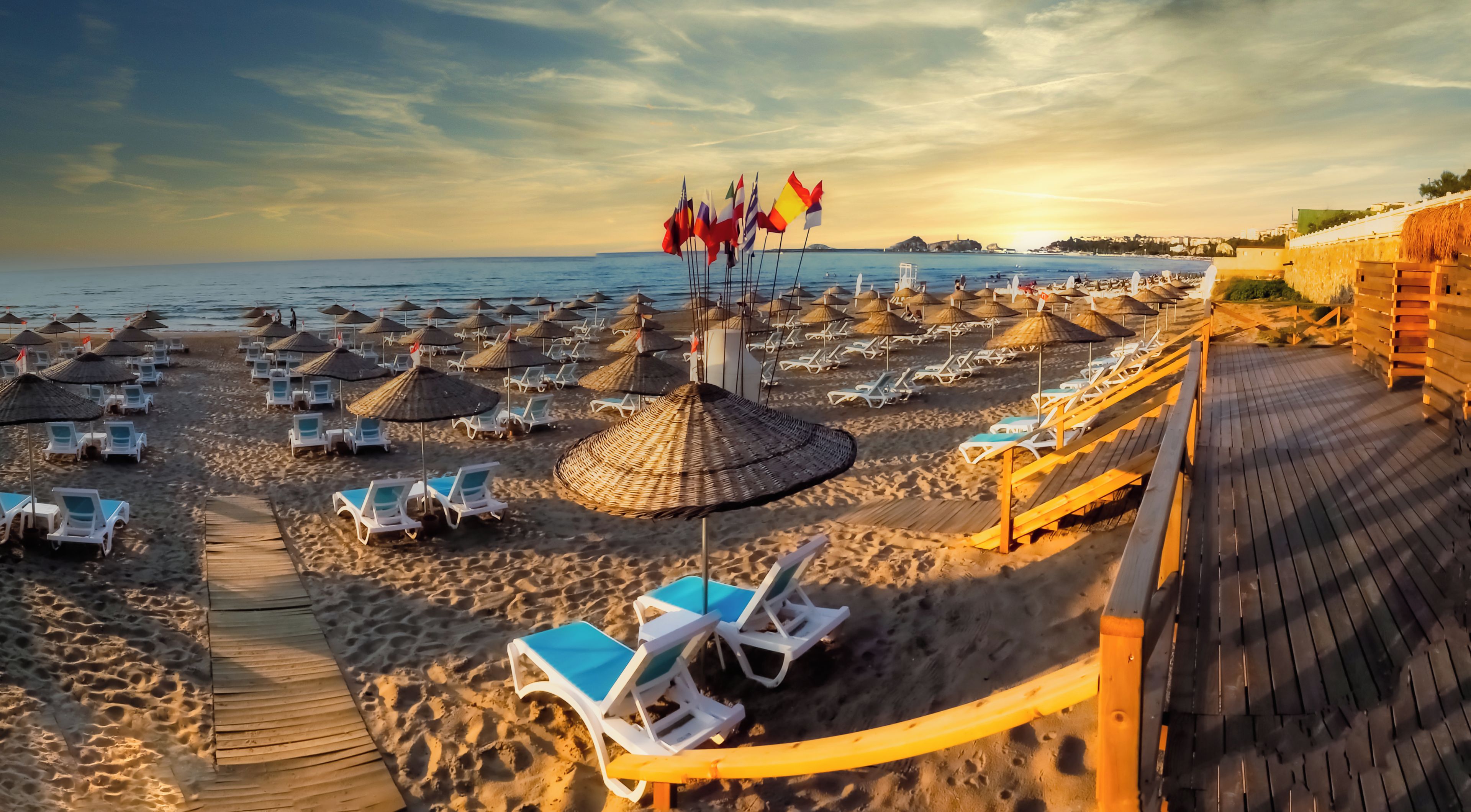 A beach with rows of sun loungers and thatched umbrellas, a wooden walkway, and flags.