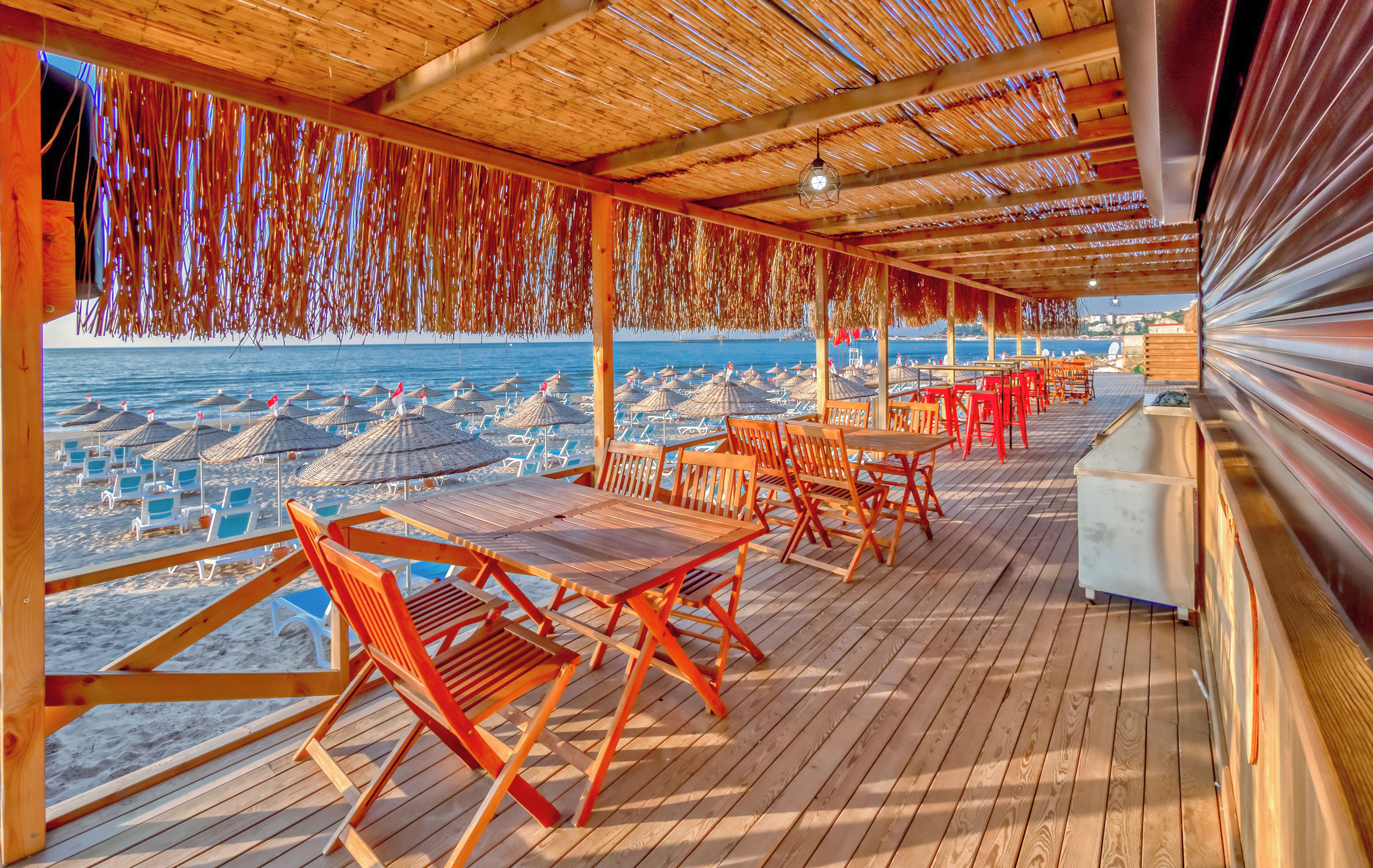 A covered outdoor seating area with wooden tables and chairs, overlooking a beach with umbrellas and lounge chairs.