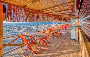 Un espacio de estar al aire libre cubierto con mesas y sillas de madera, con vistas a una playa con sombrillas y sillas de descanso.