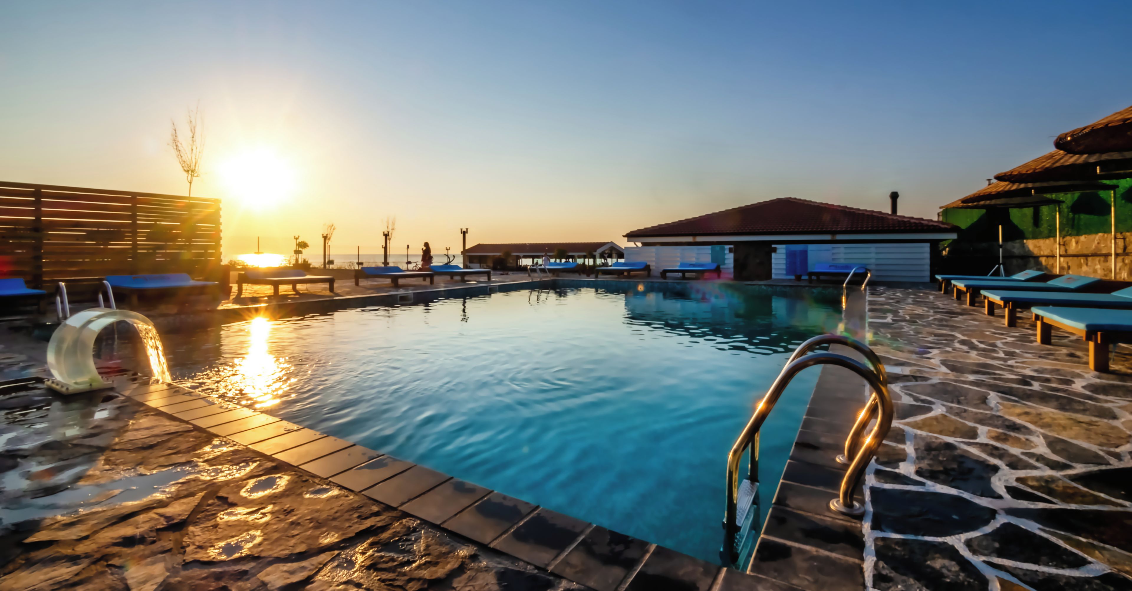 A swimming pool with a stone deck, lounge chairs, and a building with a tiled roof.