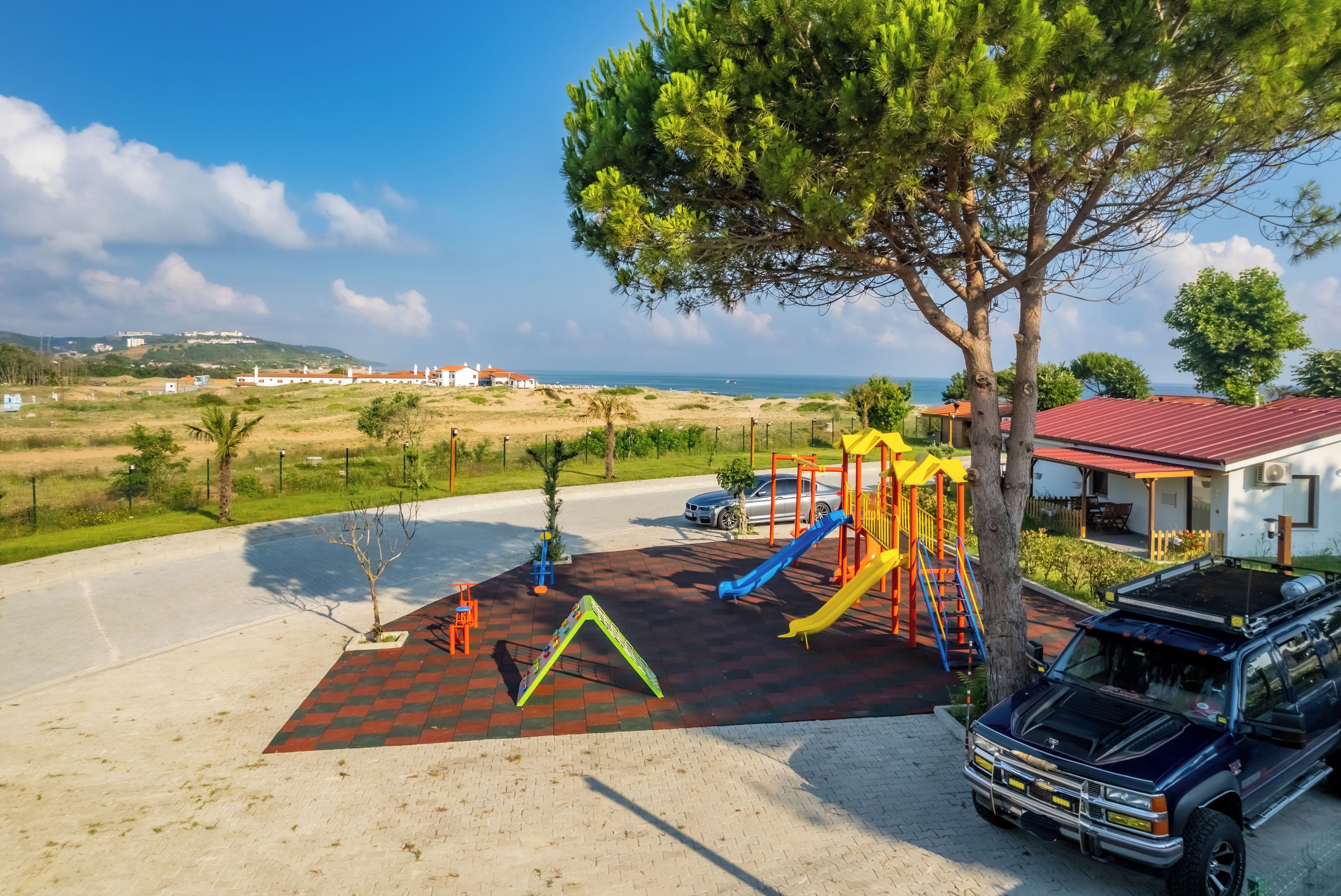 A playground with slides and climbing structures, a parking area with a vehicle, and a view of the sea and buildings in the distance.