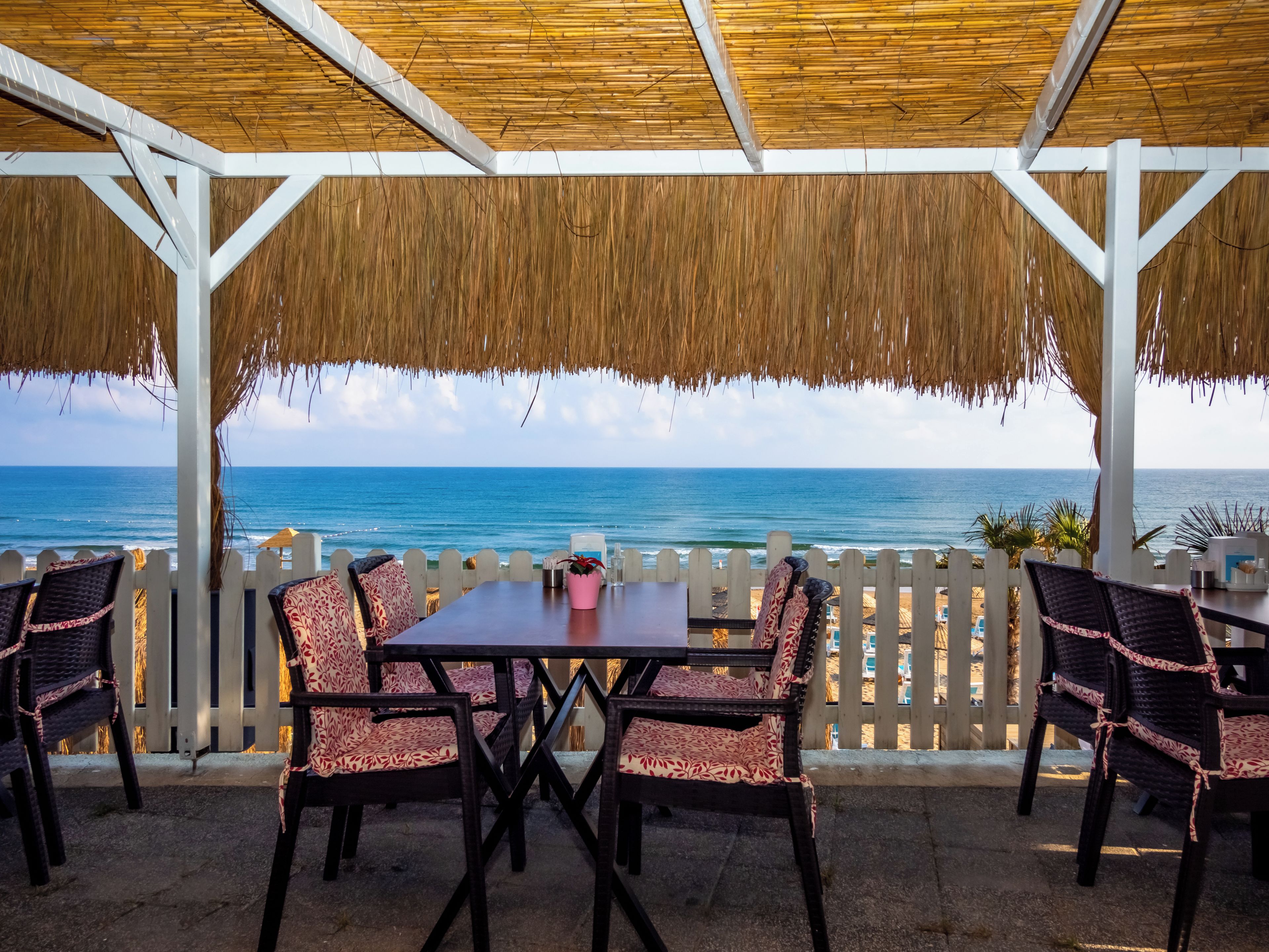 An outdoor seating area with a view of the ocean, a thatched roof, and a white picket fence.