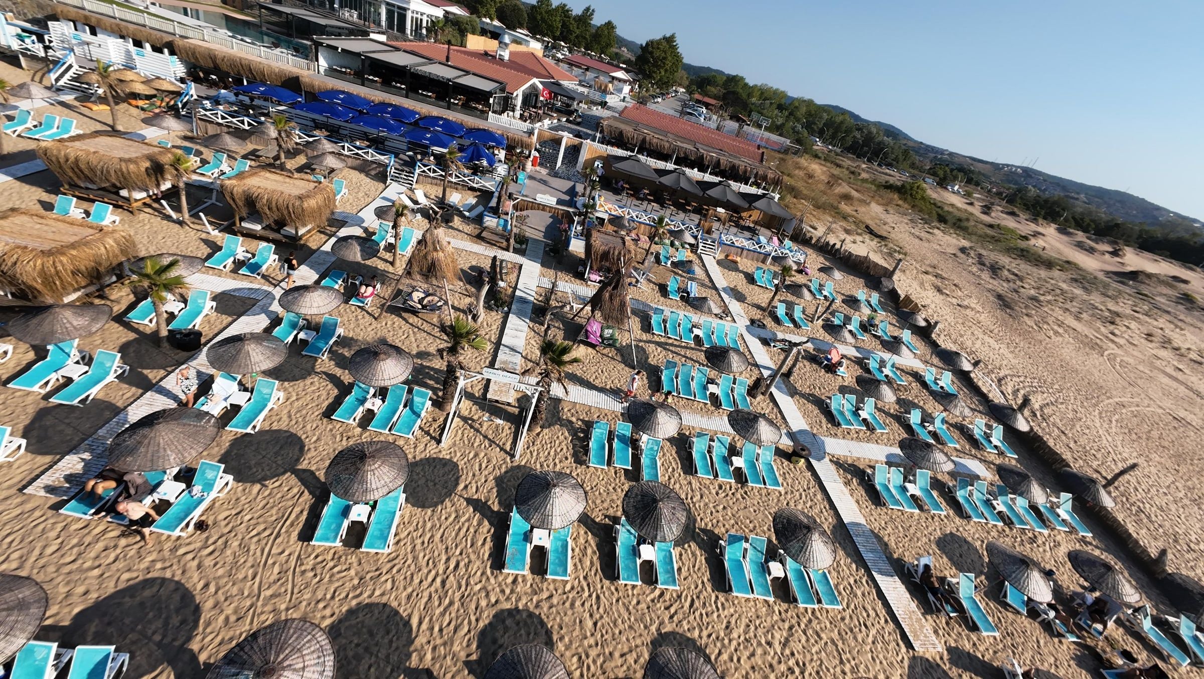 Una playa con filas de tumbonas y sombrillas, un muelle de madera y vistas del pueblo al fondo.
