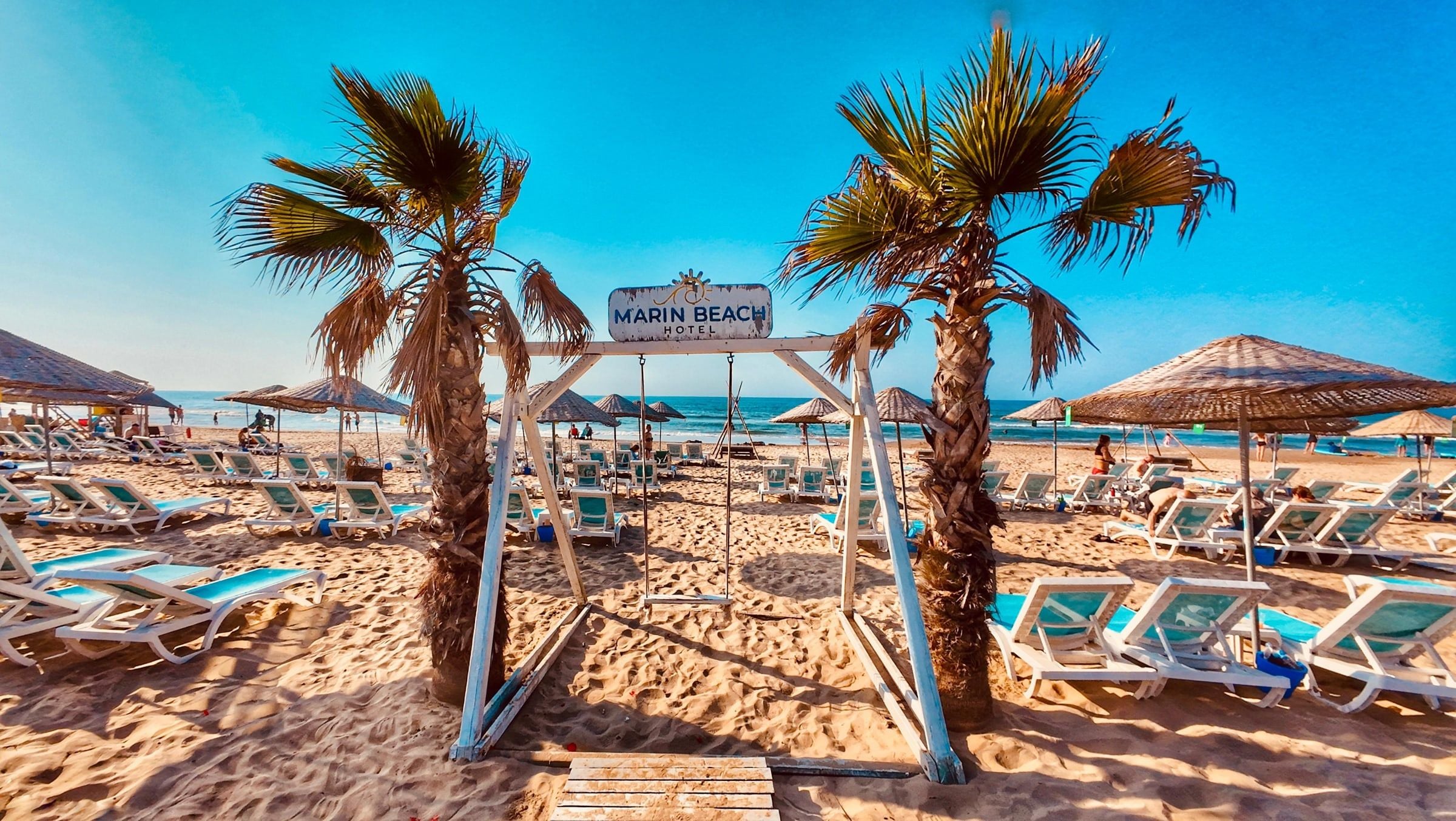 Una playa con sillas de descanso, palmeras y un cartel que indica 'Marin Beach Hotel'.